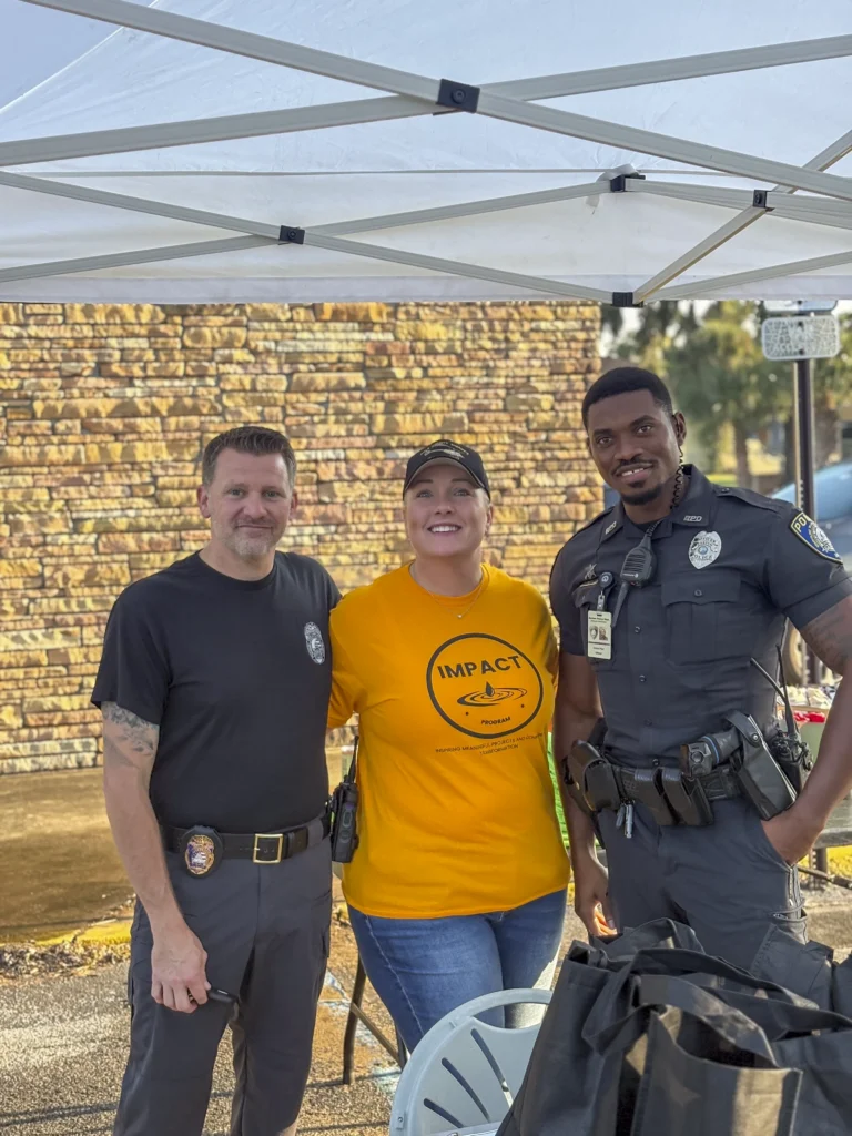 Two police officers and an Impact Program volunteer in an orange shirt posing under a white canopy tent