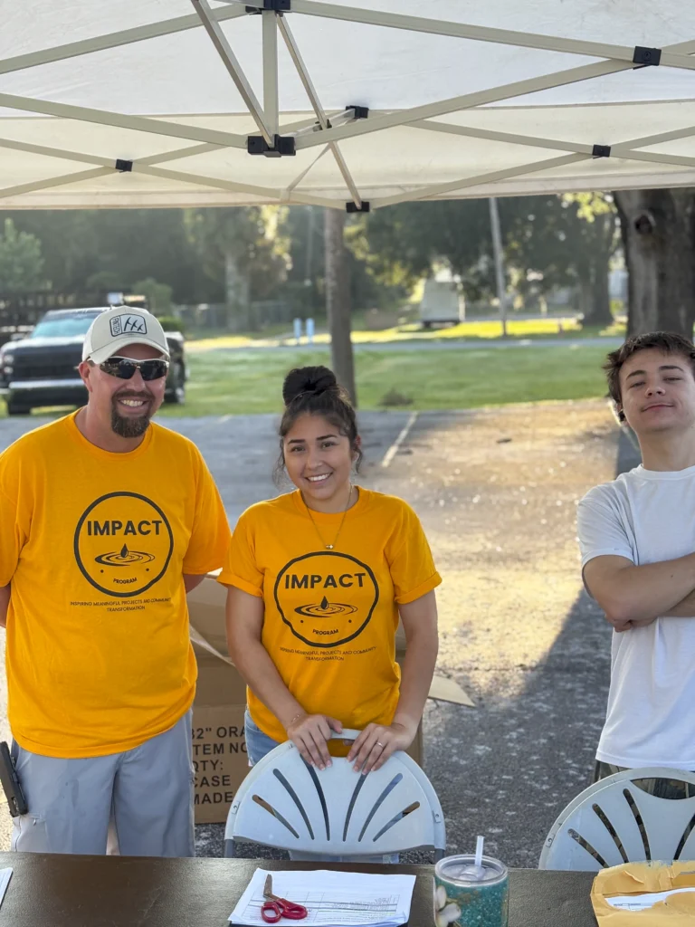 Three Impact Program volunteers in yellow shirts at a table under a white canopy tent at a Bartow CRA event