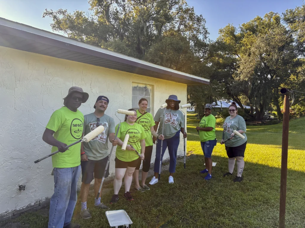 Group of Impact volunteers holding paint rollers in front of a Bartow home being painted during a CRA community project