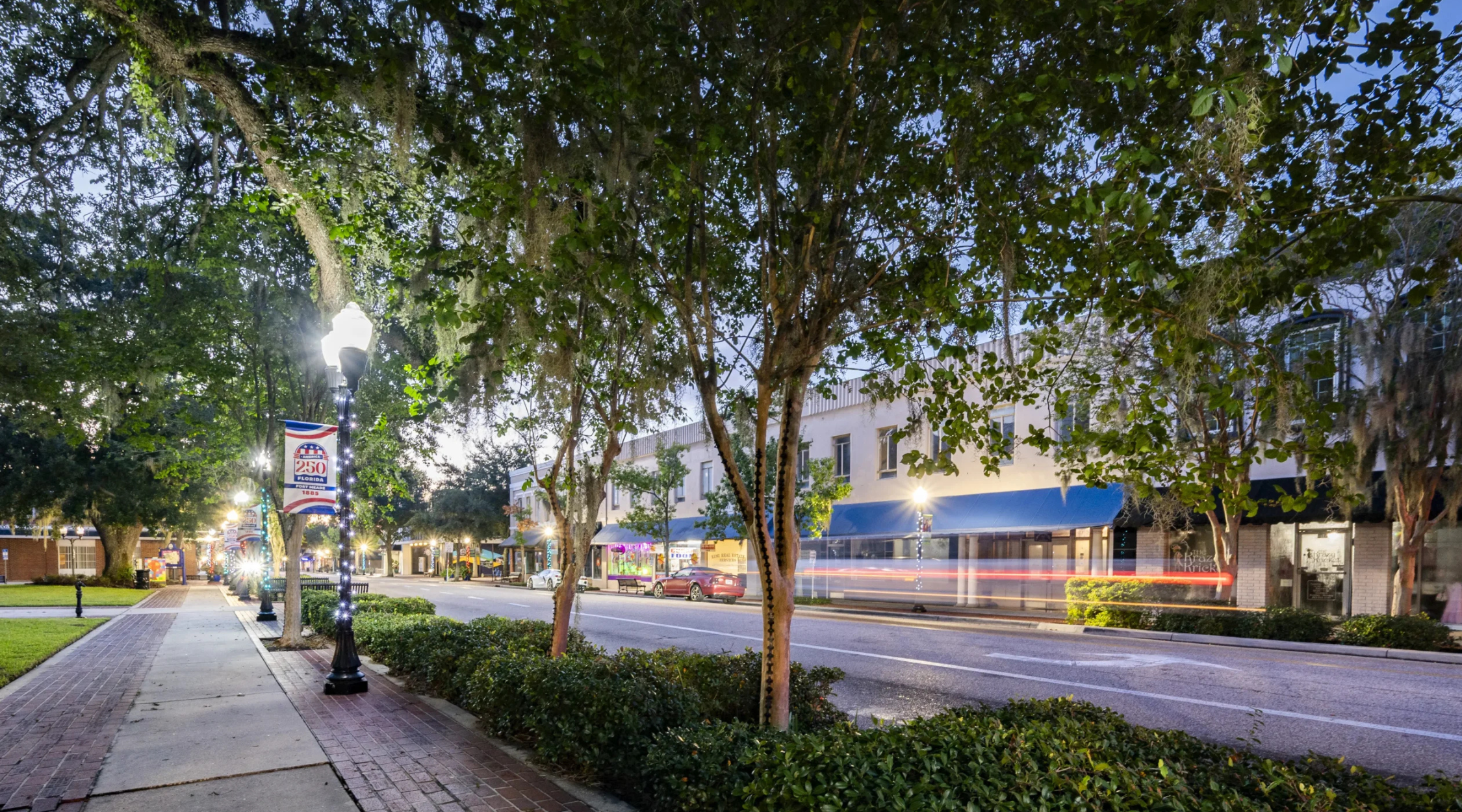 Twilight long-exposure photo of downtown Bartow's Main Street with an oak-lined sidewalk, a Florida 250 pole banner, and light trails from passing traffic