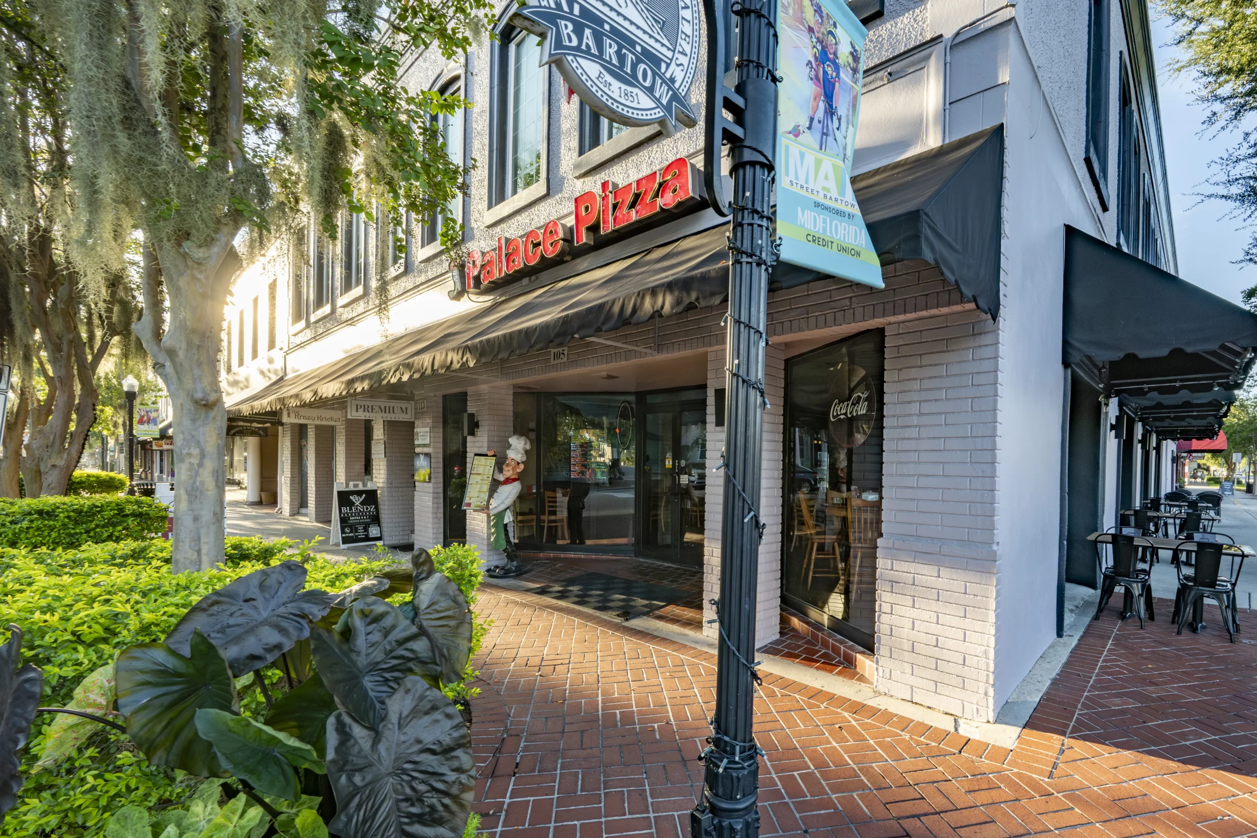 Downtown Bartow block with the Palace Pizza storefront, tropical landscaping, and oak trees along Main Street