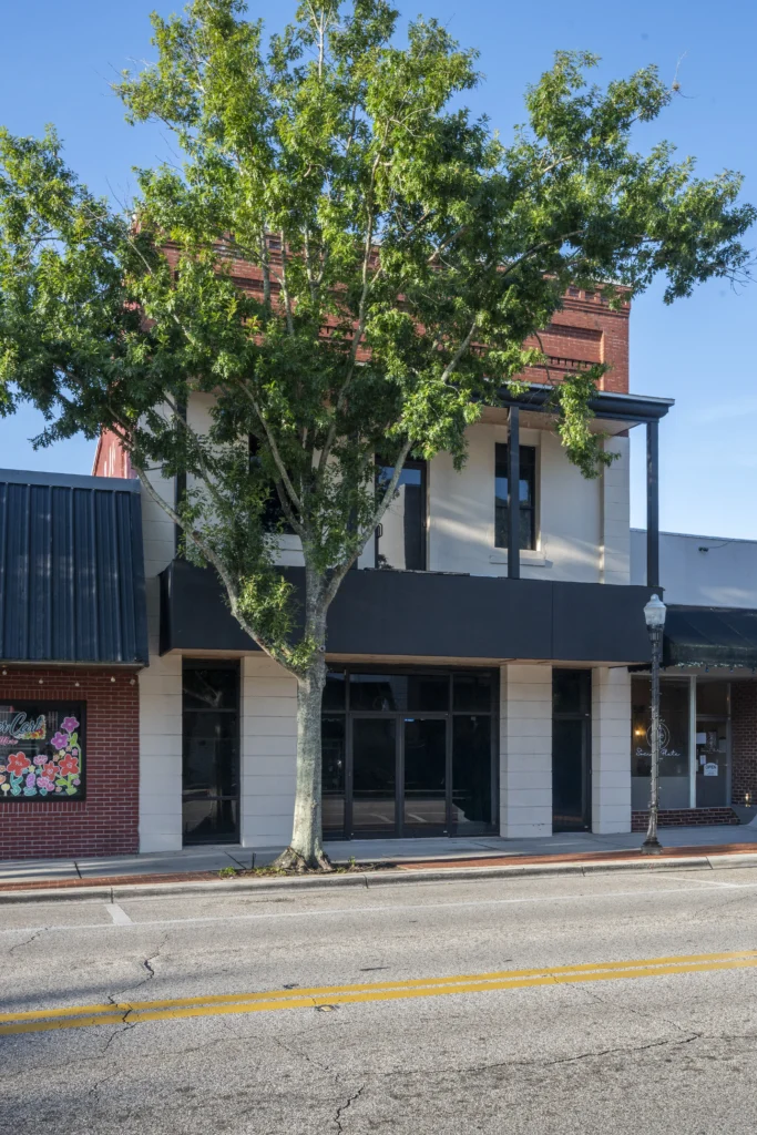 Three-story downtown Bartow building with a dark upper facade, black storefront awnings, and a large oak tree in front